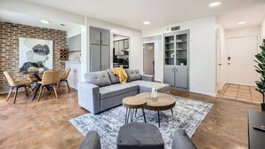 A modern living room with a grey sectional sofa, a round coffee table, and a dining area with a brick wall and a framed artwork.