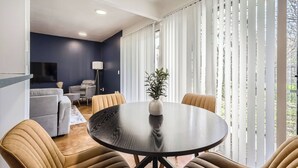 A modern living room with a round dining table, beige chairs, a grey sofa, and a TV mounted on the wall.
