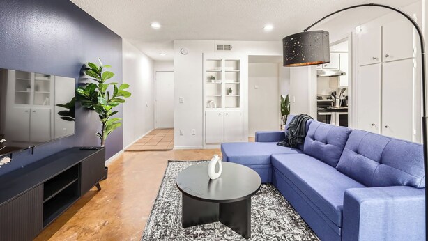 A modern living room with a blue sofa, a black coffee table, and a large mirror. There is a plant on the left and a kitchen area in the background. - Comfy King Suite Getaway | Sleeps 4 | Pool, Prkg (Austin)