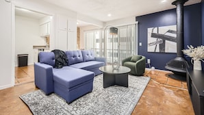 A modern living room with a blue sectional sofa, a round coffee table, and a green armchair. A black fireplace is visible on the right, and a kitchen area is in the background. - Comfy King Suite Getaway | Sleeps 4 | Pool, Prkg (Austin)