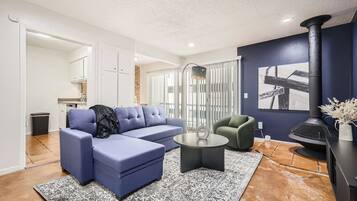 Un salon moderne avec un canapé d’angle bleu, une table basse ronde et un fauteuil vert. Une cheminée noire est visible sur la droite, et un coin cuisine se trouve au fond de la pièce.