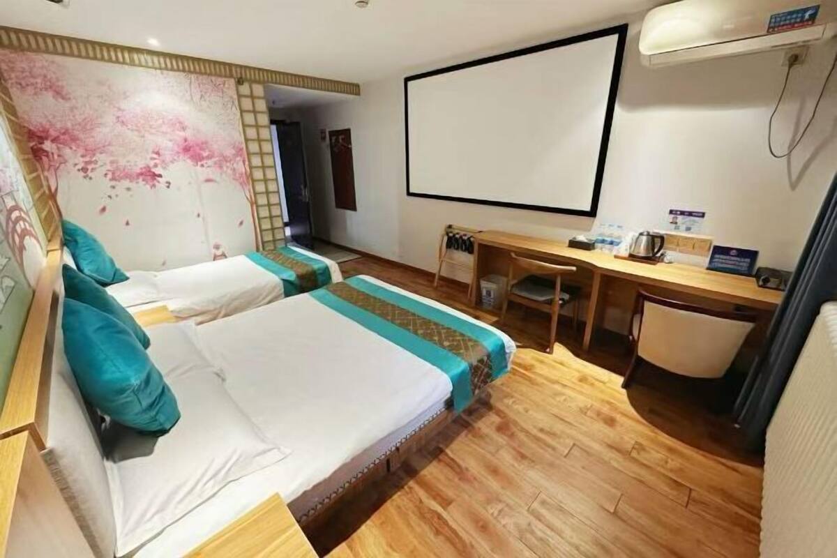 A hotel room with two beds, a desk, a chair, a large window, and a wall-mounted air conditioner.