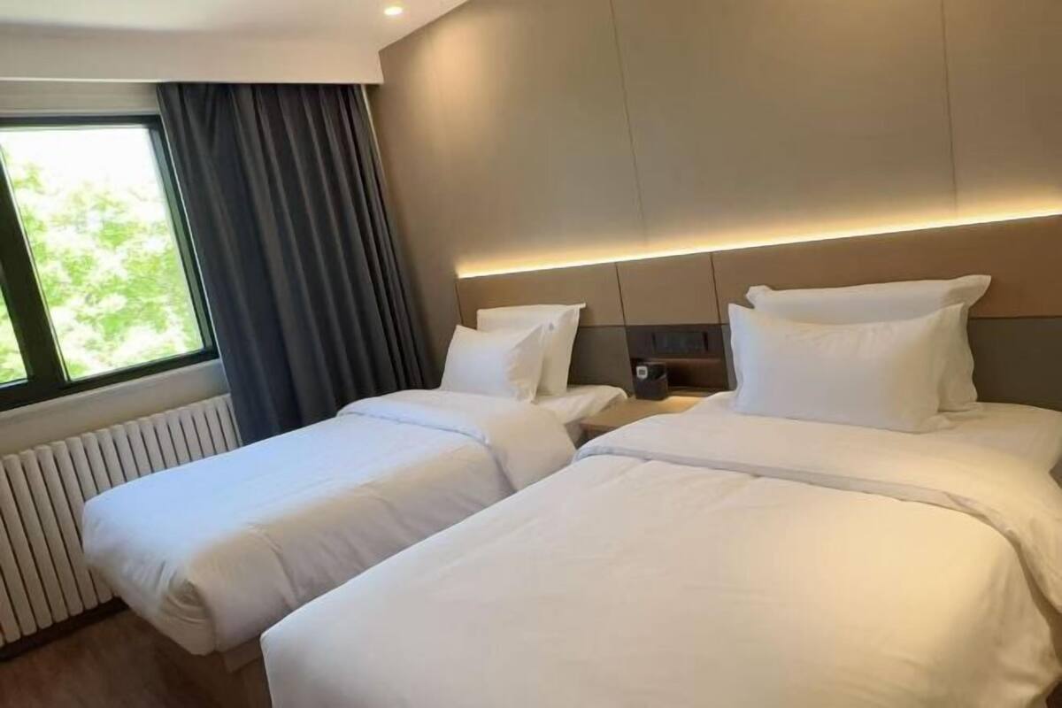 A hotel room with two beds, a window with curtains, a radiator, and a wall-mounted nightstand with a lamp.