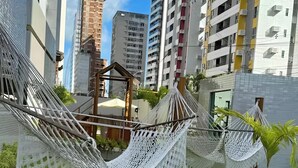 A hammock area with a small wooden structure, surrounded by a grassy lawn and a palm tree, with tall buildings in the background. - Flat in Boa Viagem (2 km from the airport) (Boa Viagem Recife)