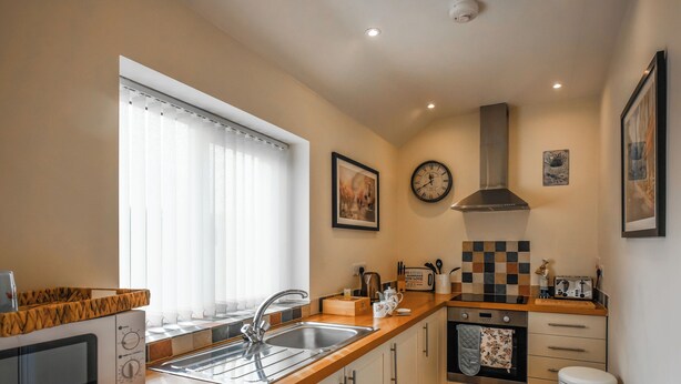 Una cocina moderna con un lavamanos, estufa y microondas. Hay una ventana con persianas, un reloj en la pared y un cubo de basura.