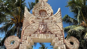 Ein türkisfarbenes Tor mit aufwendigen Schnitzereien und einem zentralen Wappen, umgeben von Palmen.