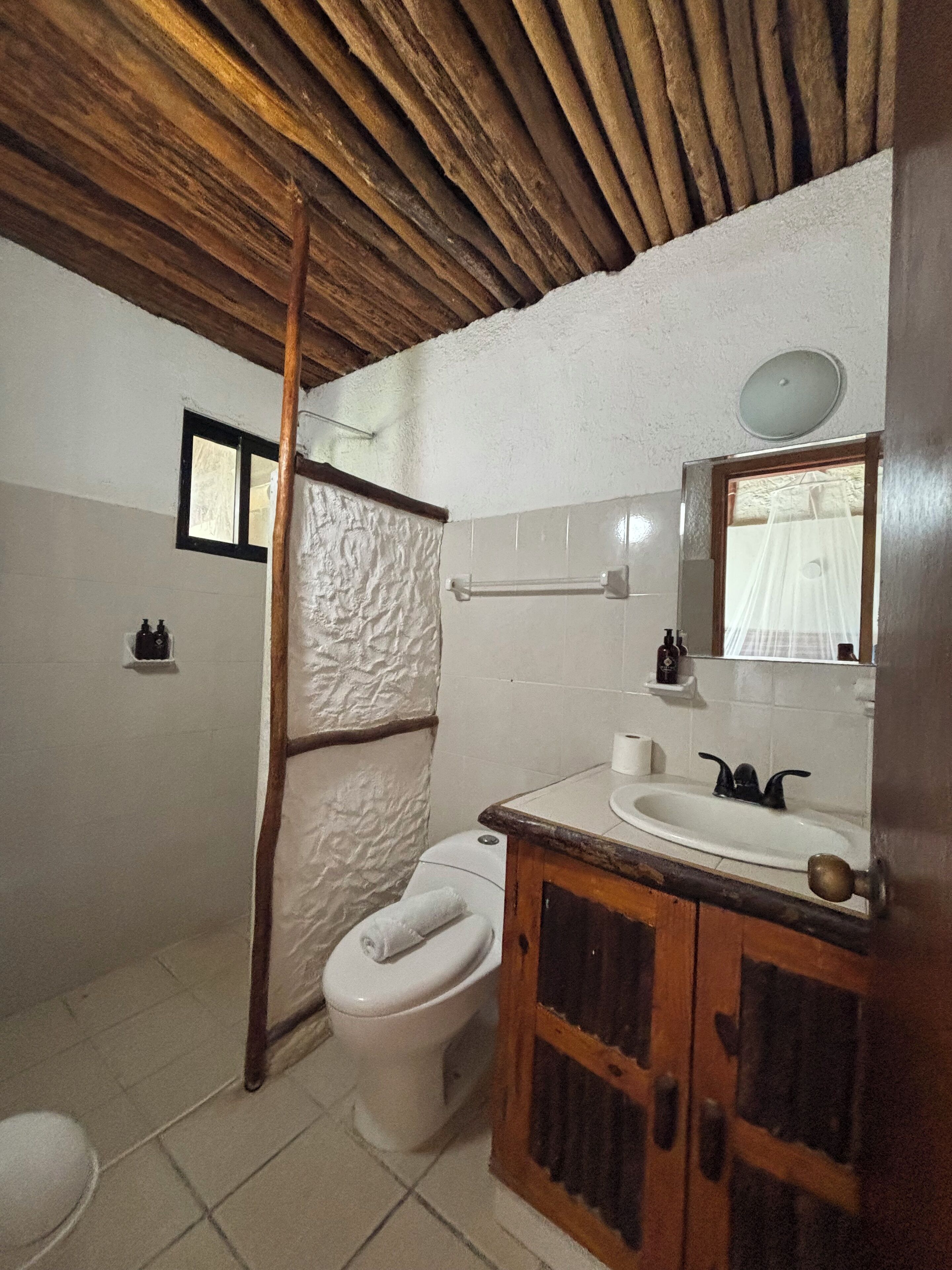 Une salle de bain avec un plafond en bois, des toilettes blanches, un lavabo avec un miroir et une serviette accrochée au mur.