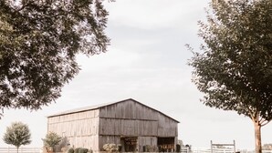 Une grande grange en bois, dotée d’un toit à pignon, entourée d’une clôture blanche et d’arbres.