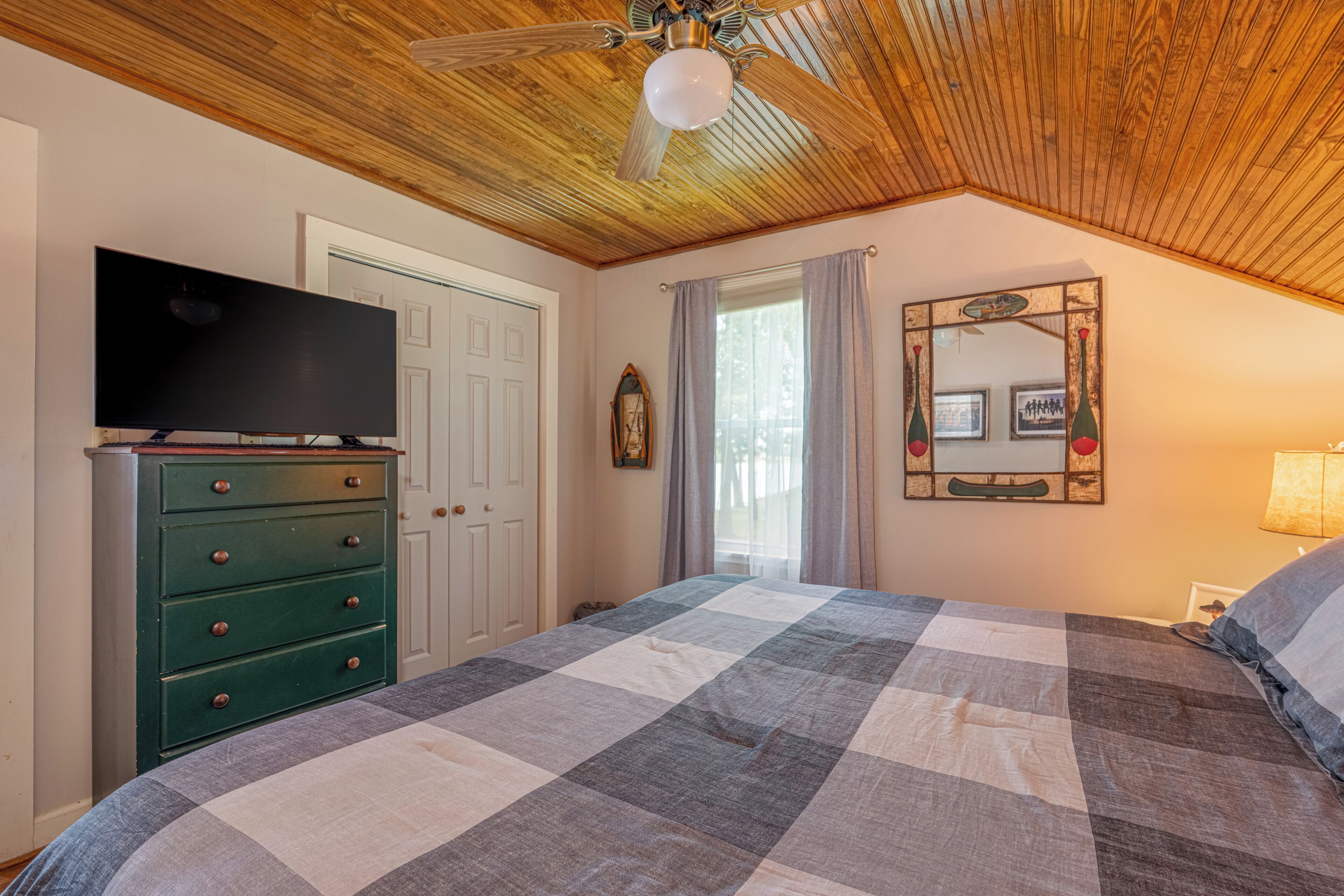 A bedroom with a wooden ceiling, a bed with a checkered quilt, a dresser with a flat-screen TV, a mirror with a decorative frame, and a window with curtains.