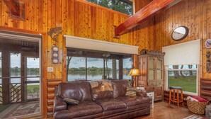 Interior de una cabaña de madera con un sofá de cuero, un ventanal con vistas a un lago y una decoración rústica.