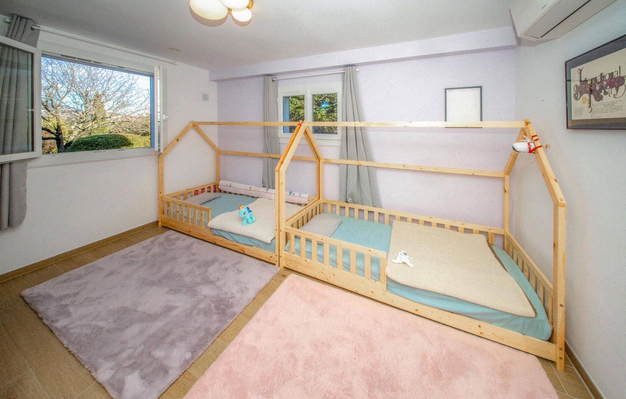 Une chambre d’enfant avec deux lits superposés, une fenêtre avec des rideaux, un tableau encadré au mur et un tapis rose sur le sol.