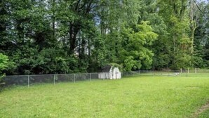 A fenced grassy area with a white structure, possibly a shed or storage unit, surrounded by dense trees.