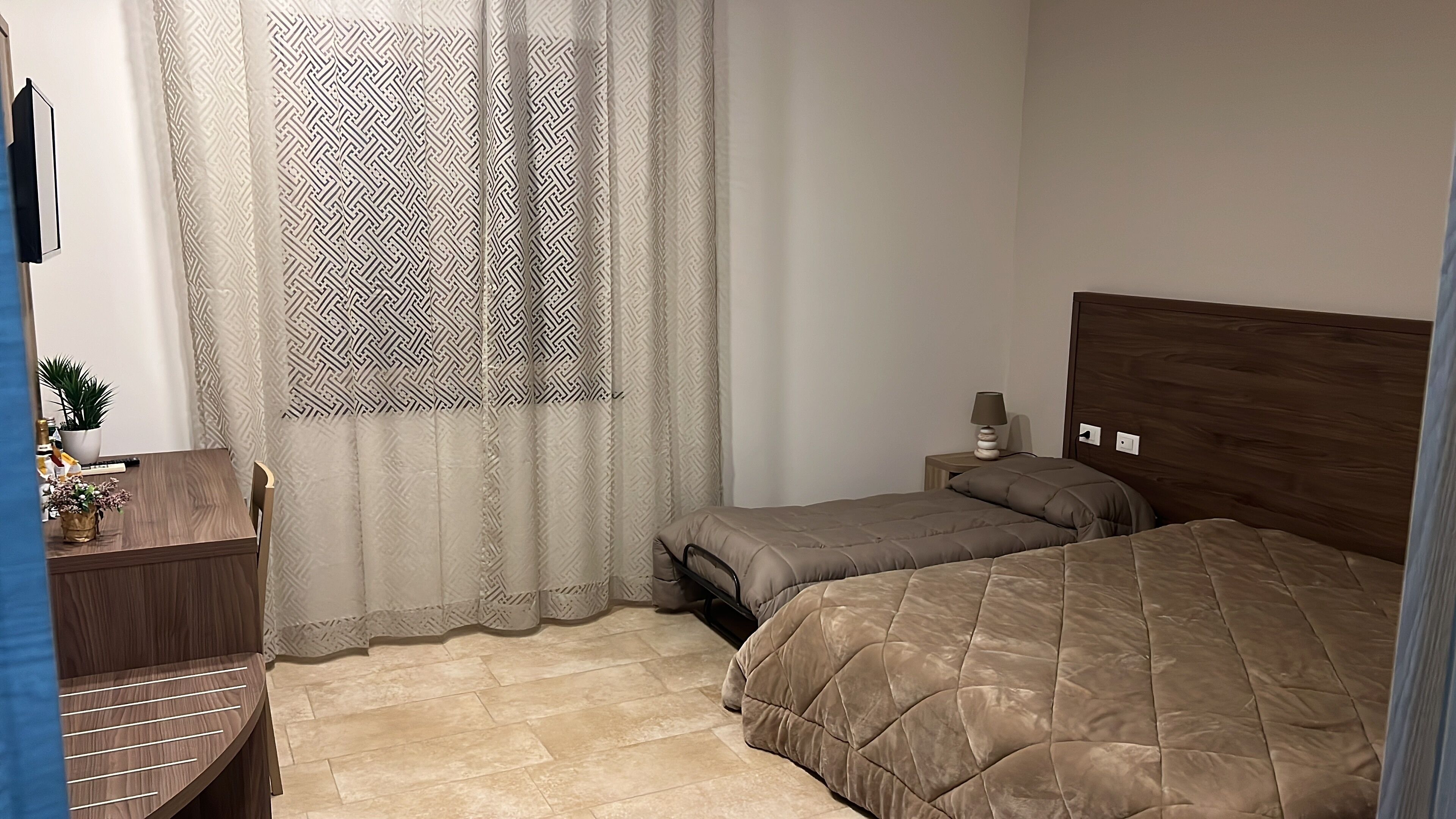 A bedroom with two beds, a wooden headboard, a bedside table with a lamp, and a window with patterned curtains.
