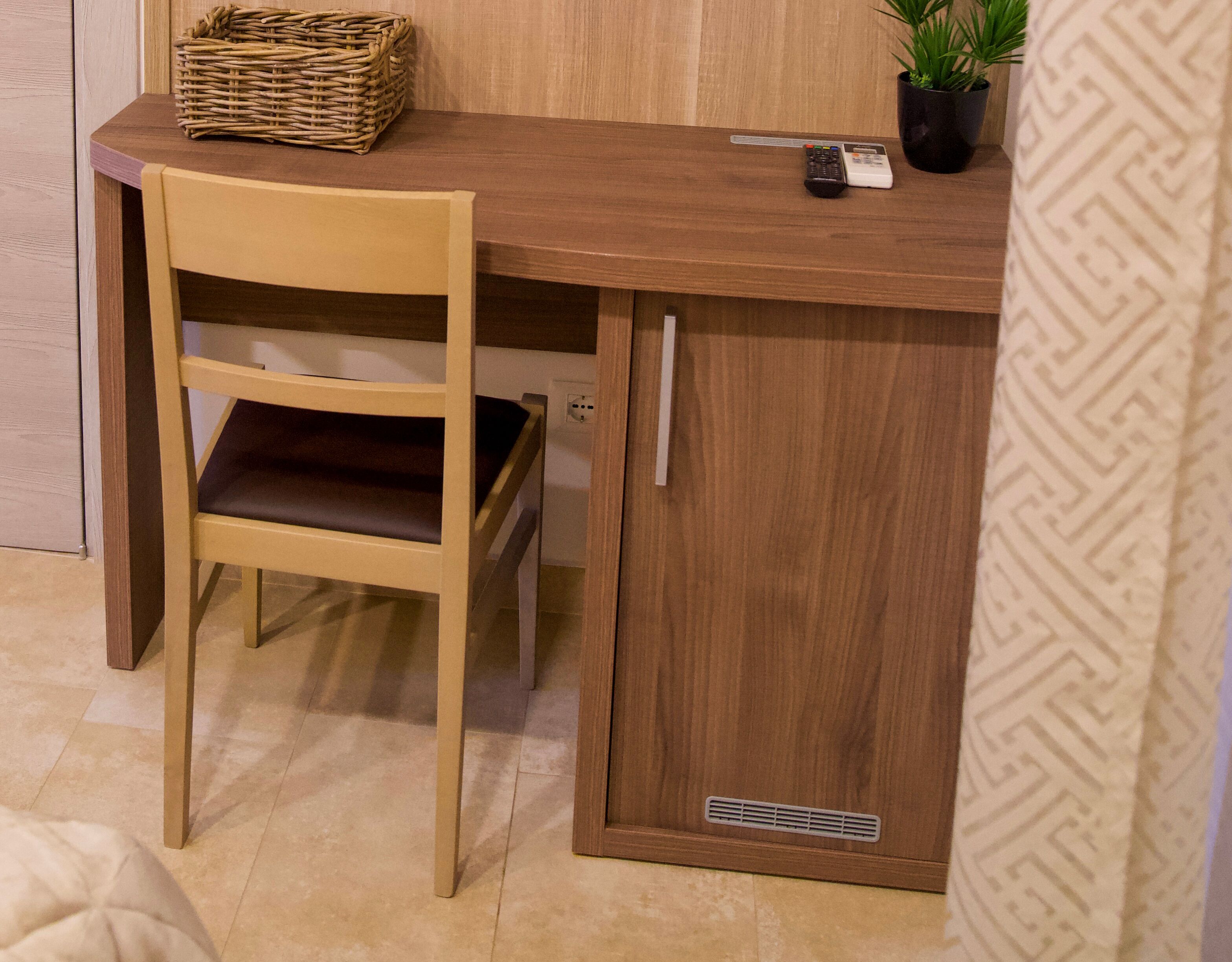A wooden desk with a chair, a basket, a remote control, and a potted plant.