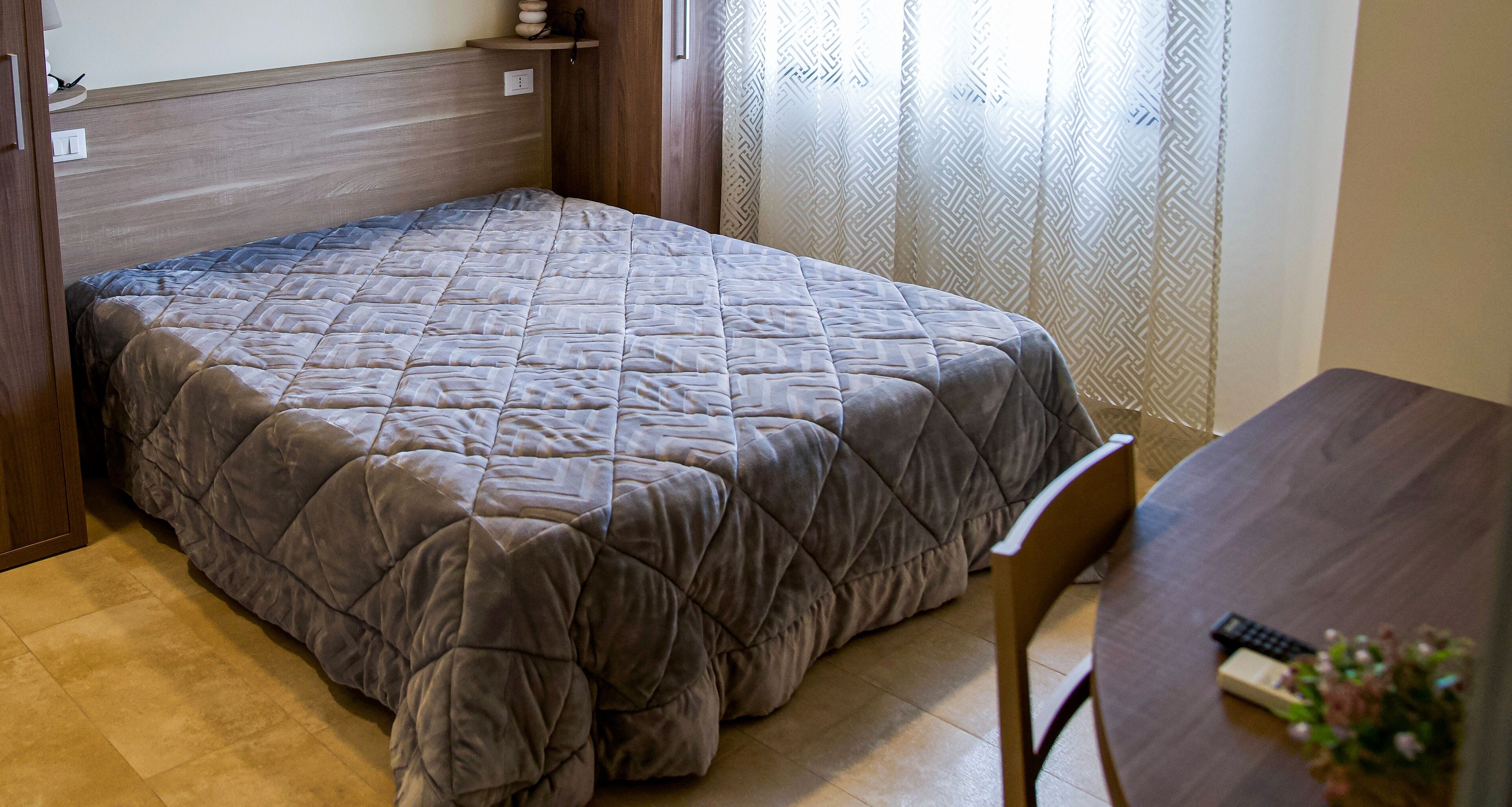 A neatly made bed with a quilted blanket, a wooden headboard, a small table with a remote control, and a chair.