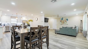 A modern kitchen with white cabinets, a dining area with wooden chairs, a grey sofa, and a TV on the wall. - Charming 3-bedroom house with AC and WiFi in awesome Kenner (Kenner)