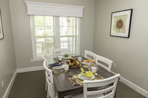 A dining room with a table set for four, white chairs, a window with blinds, and a framed picture on the wall.