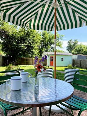 Une table ronde avec un plateau en verre, deux pichets blancs et un vase rempli de fleurs, le tout placé sous un parasol à rayures.