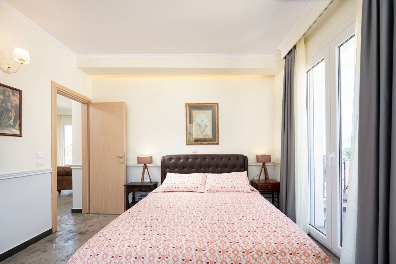 Een slaapkamer met een groot bed, een deur naar een andere kamer, een ingelijste foto aan de muur en een raam met gordijnen.