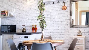Una cocina compacta con pared de ladrillo blanco, una mesa redonda de comedor con cuatro sillas, una lavadora, un microondas y un pequeño estante con objetos decorativos.