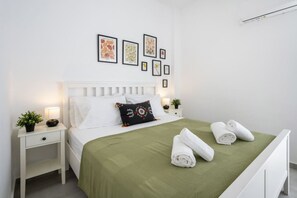 A neatly arranged bedroom with a white bed, green bedspread, and white towels on the bed. There are two bedside tables with lamps and small potted plants. The walls are adorned with framed botanical prints.
