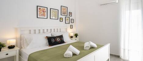 A neatly arranged bedroom with a bed, a bedside table, a lamp, a wall-mounted air conditioner, and framed pictures on the wall.