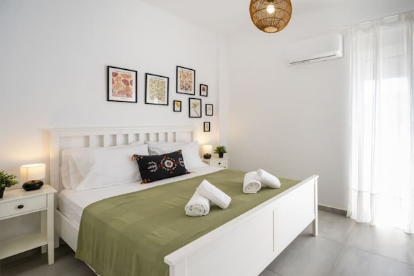 A neatly arranged bedroom with a bed, a bedside table, a lamp, a wall-mounted air conditioner, and framed pictures on the wall.