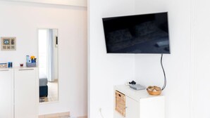 A modern, minimalist living room with a flat-screen TV mounted on the wall, a white cabinet with a basket of bread, and a large mirror reflecting the room. - Sunrise Beachfront Getaway (Kardamaina)