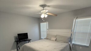 A bedroom with a bed, a ceiling fan, a TV on a stand, and windows with blinds.