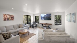 Un vaste salon comprenant un canapé, une table basse et un coin repas avec des chaises et une table. De grandes fenêtres laissent entrer la lumière naturelle et offrent une vue sur l’extérieur.
