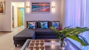 A modern living room with a dark sofa, decorative pillows, a glass coffee table, and a painting of a sunset over the ocean. - Cozy 2 bethoom apartament (Andrés)