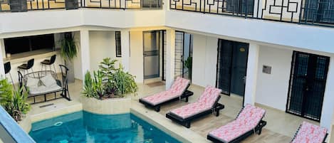 Une piscine avec des chaises longues, un toit de chaume et un bâtiment à plusieurs niveaux doté de balcons.