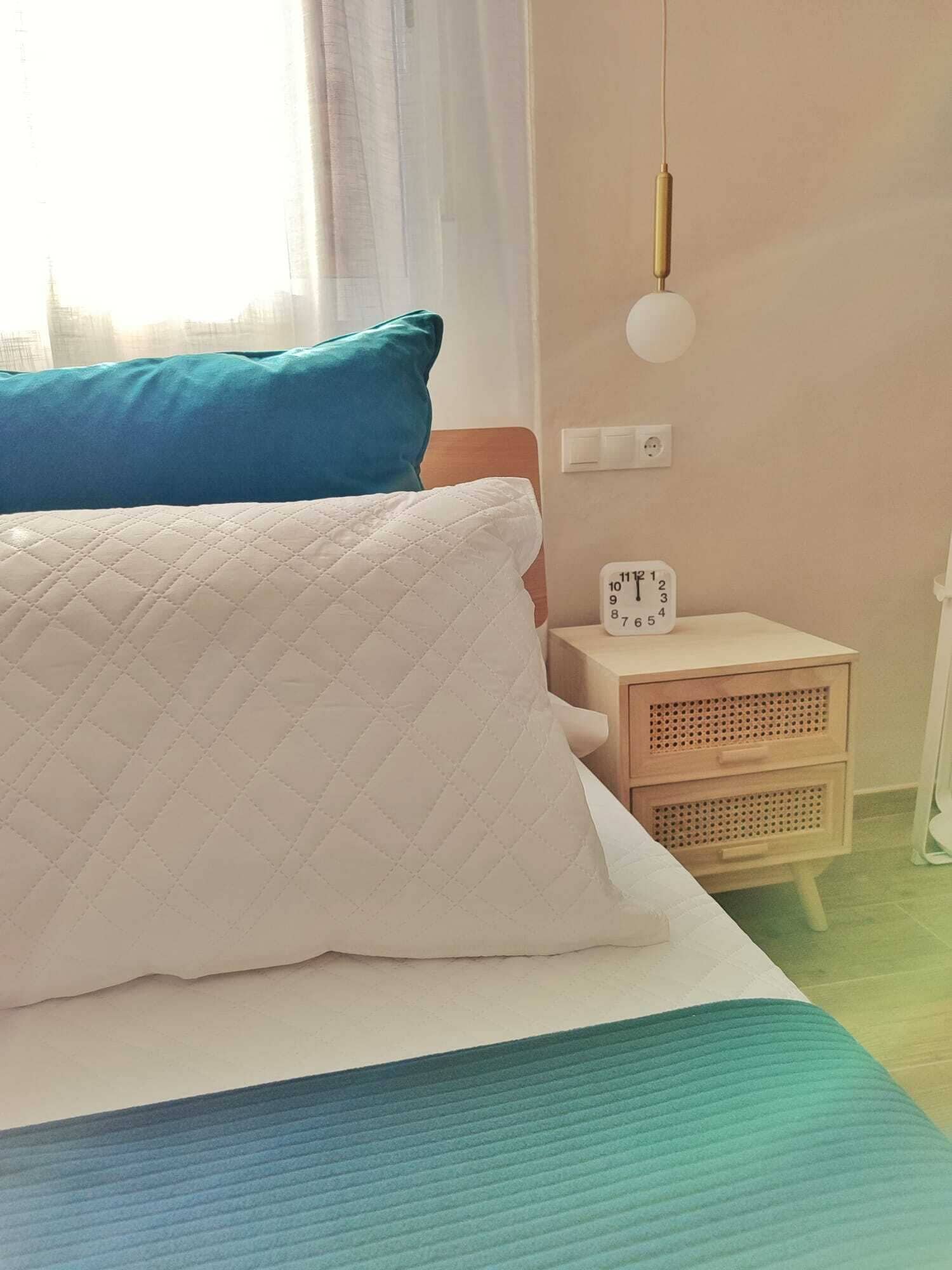 A bed with a quilted white pillow and a teal blanket, a bedside table with a clock, and a hanging light fixture.