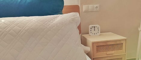 Una cama con un almohadón blanco acolchado y una manta color turquesa, una mesita de noche con un reloj, y una lámpara colgante.