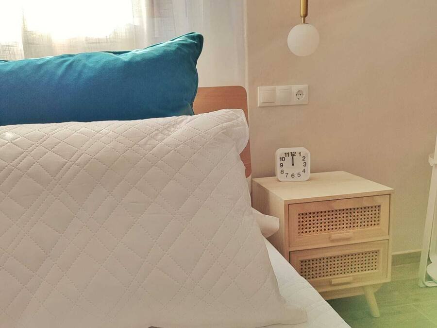 A bed with a quilted white pillow and a teal blanket, a bedside table with a clock, and a hanging light fixture.