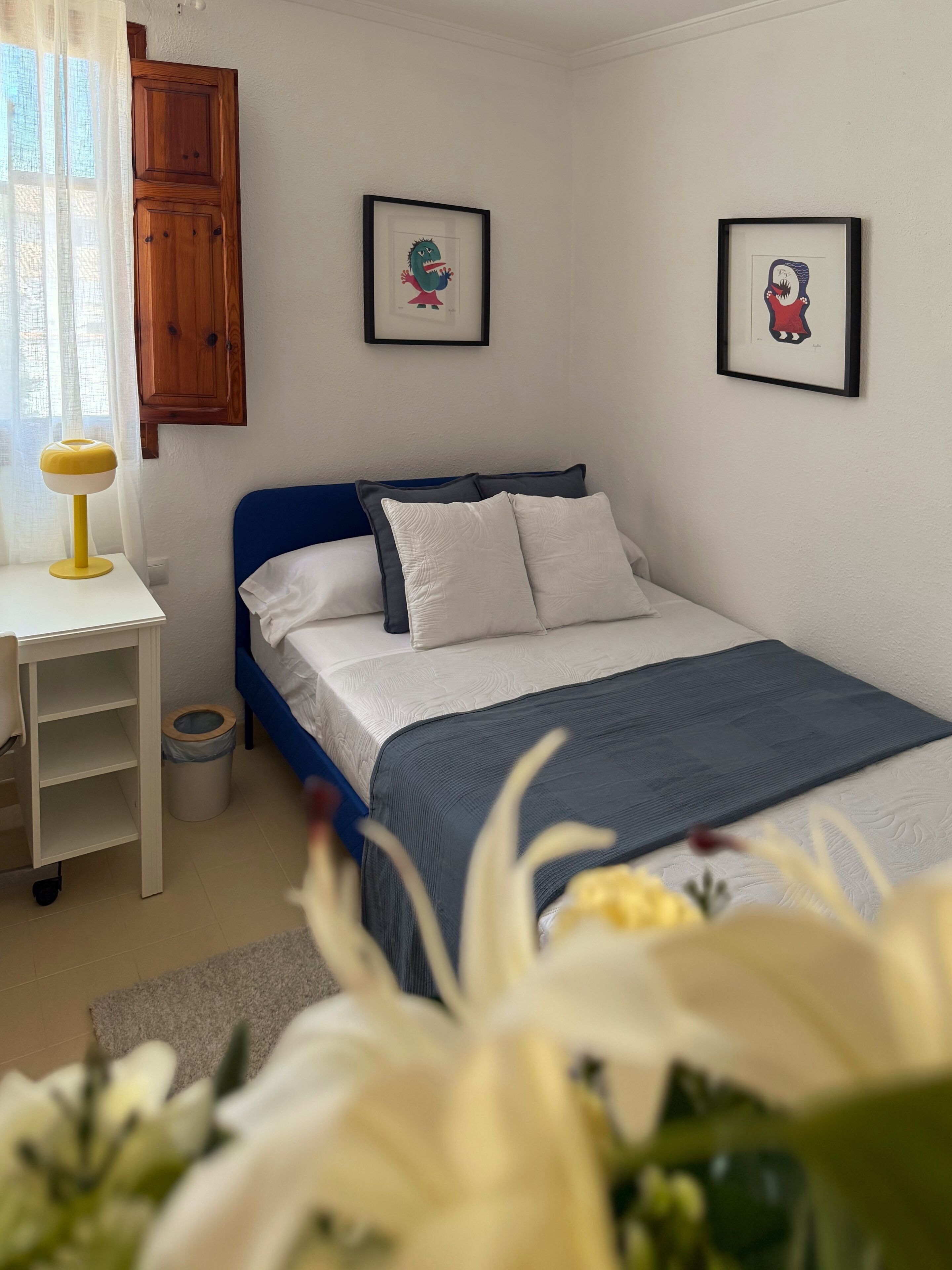 Un dormitorio con una cama, un escritorio, una silla, un bote de basura y dos cuadros en la pared.