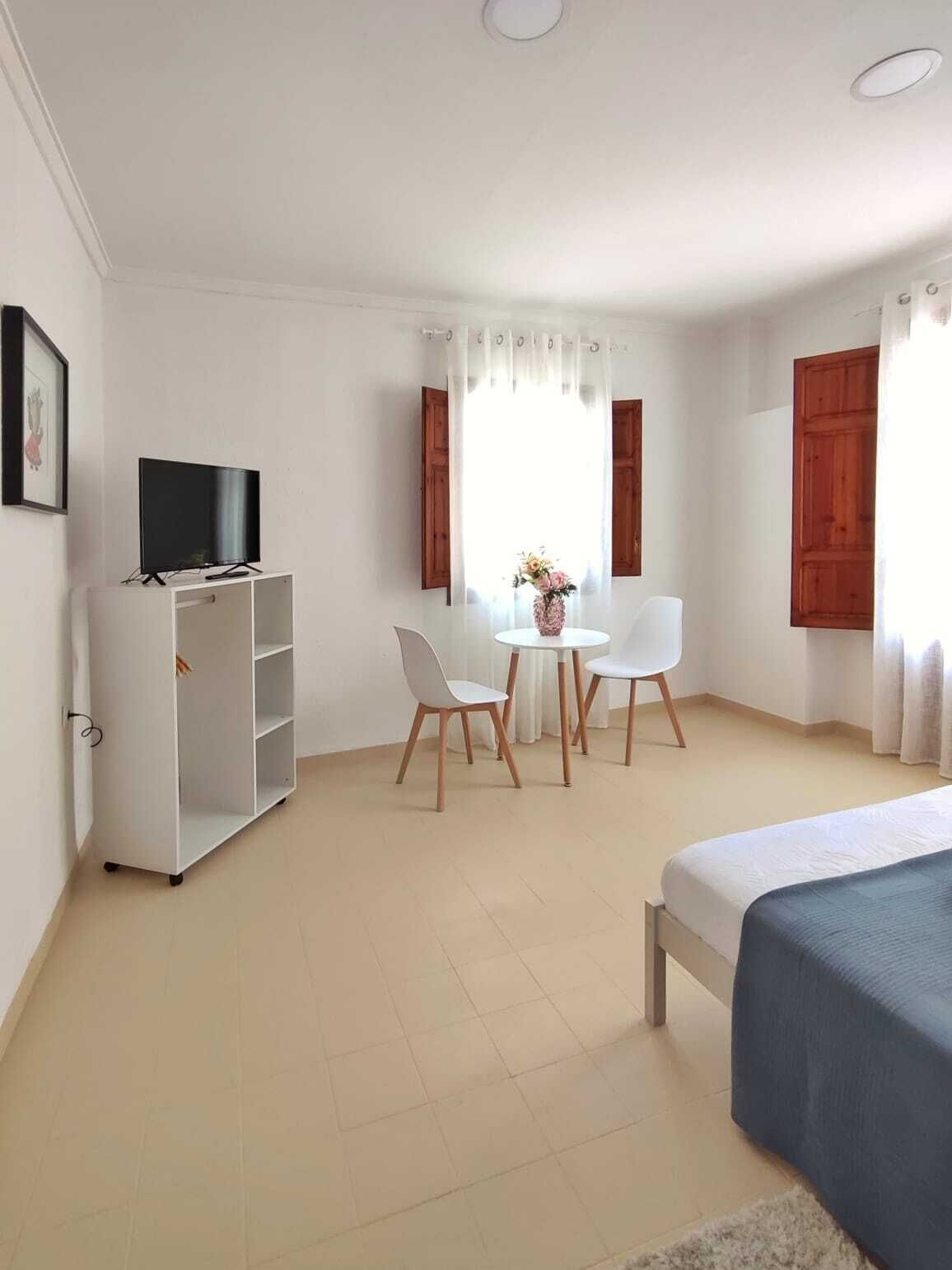 Un dormitorio con cama, televisor sobre un mueble blanco, mesa de comedor con sillas y ventana con cortinas.