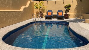 En swimmingpool med flisekant, to liggestole med orange puder og plantekrukker.