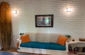 Un mur en briques blanches orné d’un tableau encadré, un canapé blanc avec des coussins turquoise et une planche de surf en bois appuyée contre le mur.