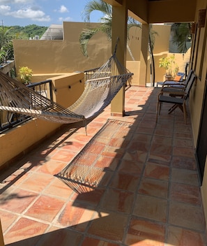 A hammock and chairs on a tiled patio with a view of a hill and trees.