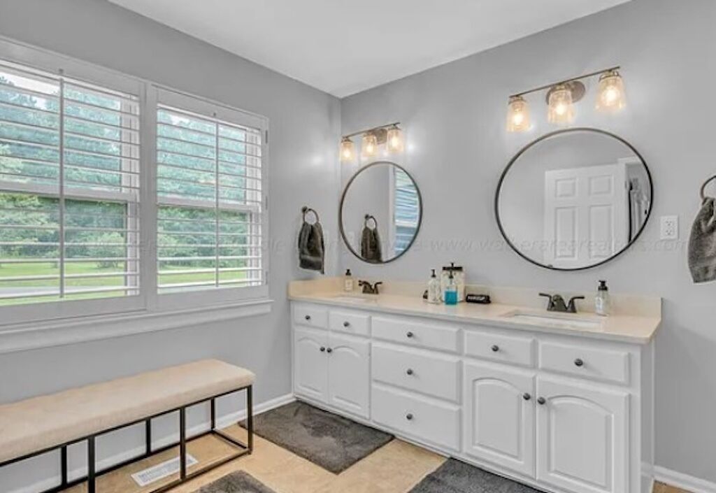 A bathroom with a vanity, two mirrors, a bench, and a window with blinds.