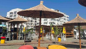 Una spiaggia con ombrelloni di paglia, lettini e un edificio bianco con balconi sullo sfondo.