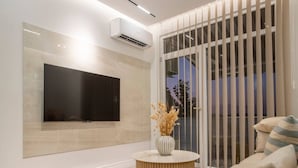 Un soggiorno moderno con una TV a schermo piatto fissata al muro, un tavolino rotondo in legno con un vaso di fiori secchi e una porta scorrevole in vetro che dà su uno spazio esterno.