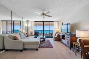 A spacious living room with a large flat-screen TV, a ceiling fan, and a view of the ocean through floor-to-ceiling windows.