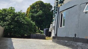 Un bâtiment à l’extérieur gris, doté d’une fenêtre et d’un petit jardin avec des plantes et un réservoir d’eau noir.