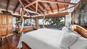 Une chambre spacieuse avec un grand lit, des poutres apparentes en bois au plafond, une armoire en bois et une vue sur les palmiers.