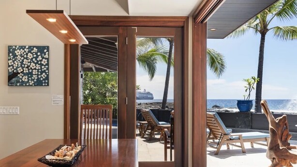 Une table de salle à manger en bois, sur laquelle se trouve un saladier rempli de collations, une chaise en bois, et une vue sur la plage avec des chaises longues et des palmiers.