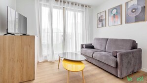 A modern living room with a grey sofa, a wooden TV stand, a yellow coffee table, and a large window with sheer curtains. - Pleasant apt with balcony -6P-Proche Disney/Paris (Champigny-sur-Marne)