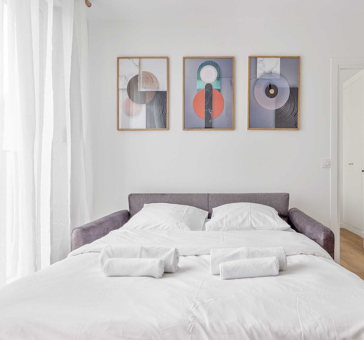 A neatly made bed with white linens and towels, a gray headboard, and three framed abstract artworks on the wall.