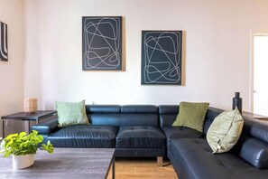 Un salon moderne avec un canapé en cuir noir, deux œuvres d’art abstraites accrochées au mur, une petite table d’appoint et une plante en pot.